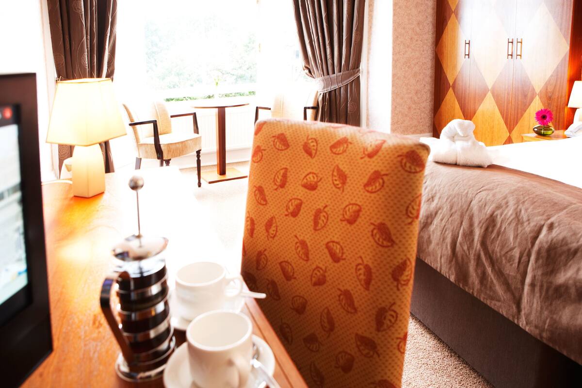 Una habitación de hotel con cama, una silla, una mesa con tazas y una cafetera, y cortinas.