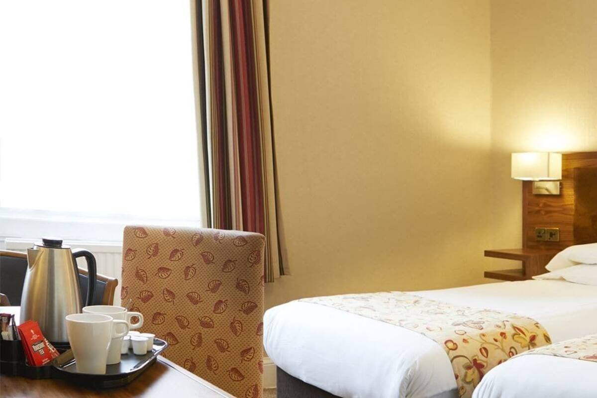 Habitación de hotel con dos camas, una silla, una bandeja con tazas de café y una ventana con cortinas.