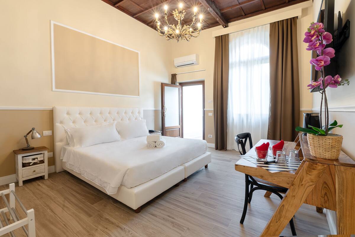 A bedroom with a large bed, a wooden dining table, a chandelier, and a potted orchid.