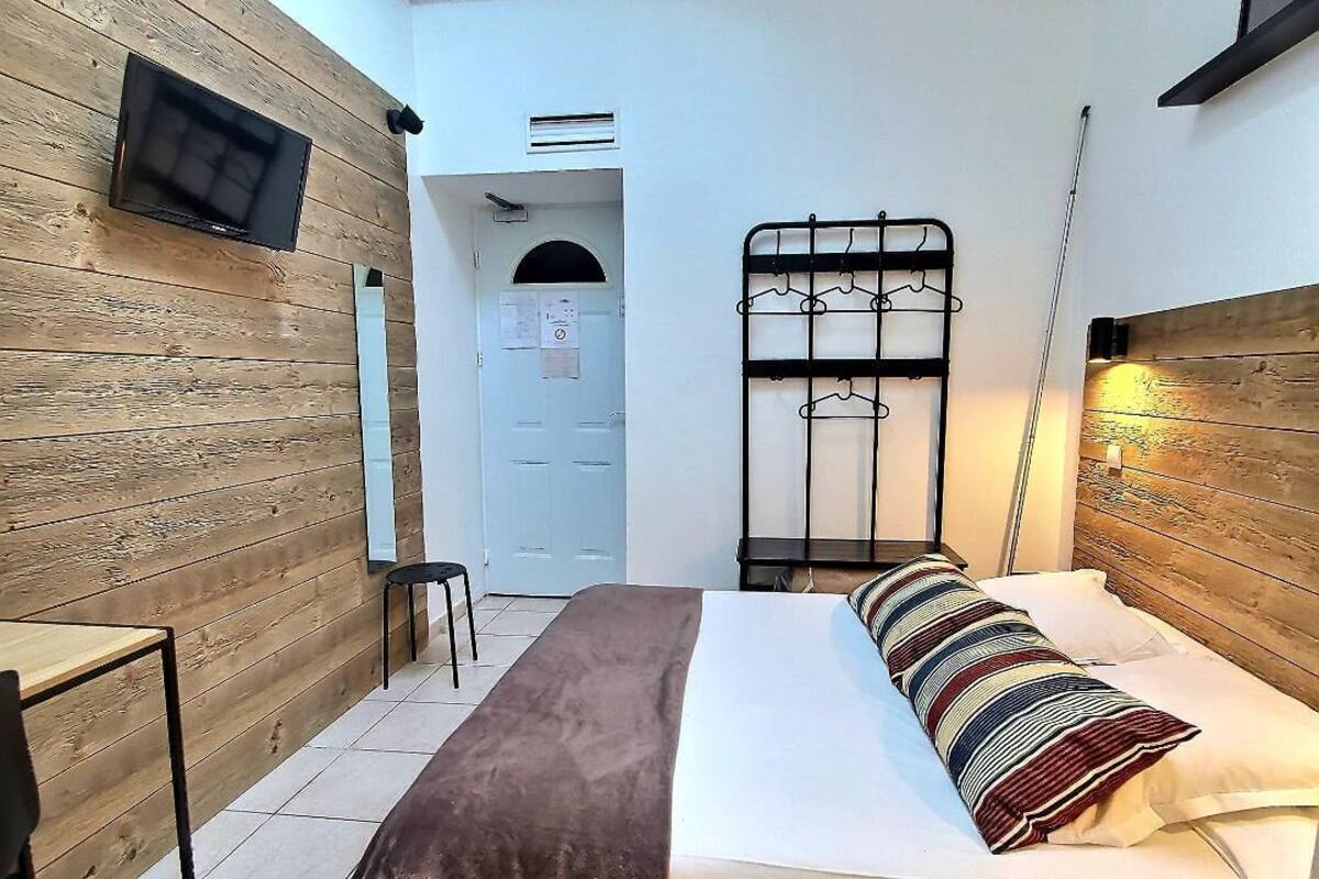 A bedroom with a bed, a TV mounted on a wooden wall, a metal rack, and a door leading to another room.