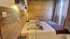 Habitación de hotel con cabecera de madera, cama con ropa de cama blanca, control remoto sobre una mesita y una ventana con cortinas.