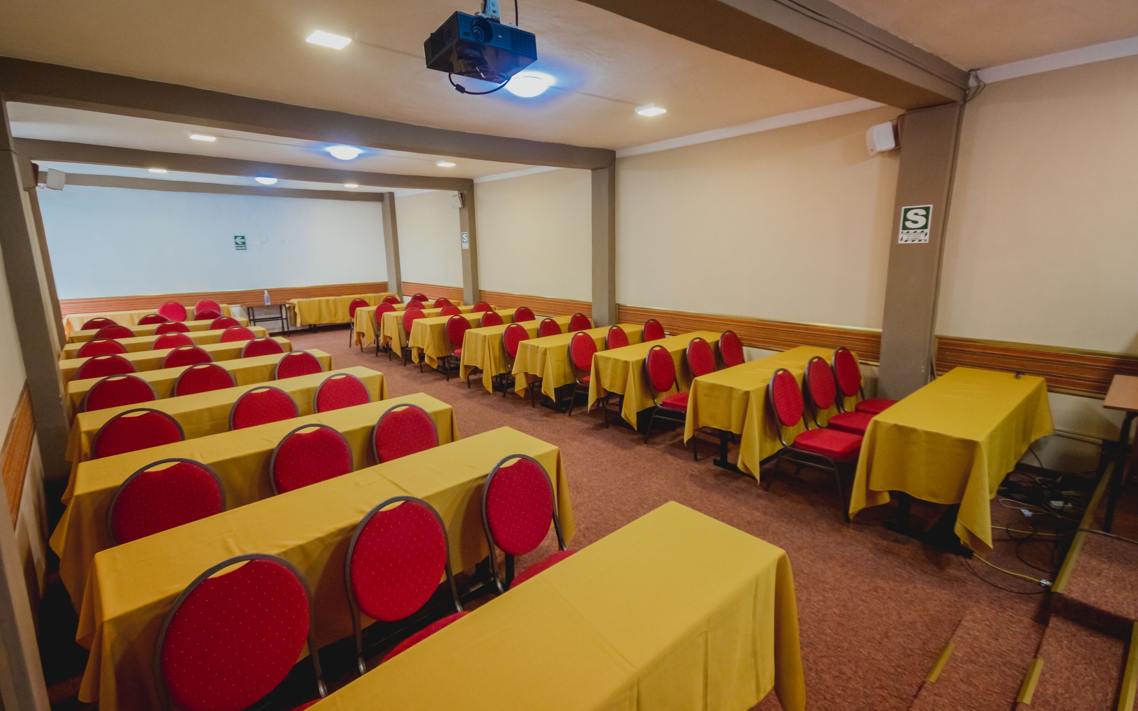 A conference room with rows of tables and chairs, equipped with a projector and ceiling lights.