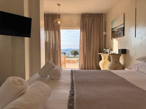 A hotel room with a bed, towels, a TV, a table with a lamp, and a view of the outdoors. - Eight Dimensions Capari Suites Elafonisos (Elafonisos)