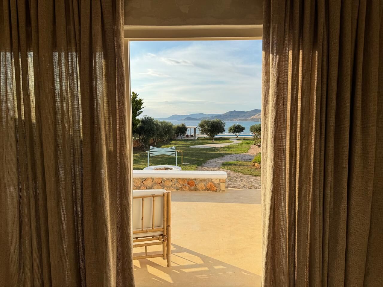 Vista a través de una ventana con cortinas, que muestra un muro de piedra, un banco y un paisaje con árboles y montañas.