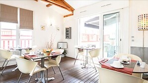 Ein modernes Hotelzimmer mit Holzbalkendecke, großen Fenstern, einer Essecke mit weißen Stühlen und Blick auf die Stadt.