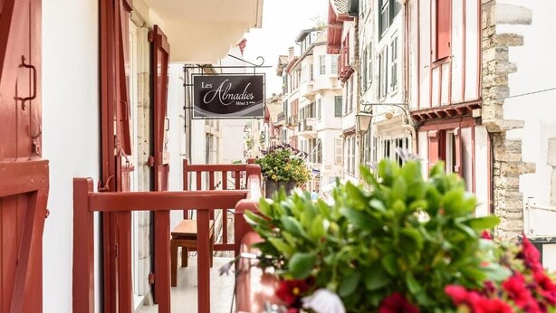 Eine enge Straße mit traditioneller europäischer Architektur, ein Schild für das Hotel 'Les Abadies' und ein Blumenbeet mit roten Blüten.