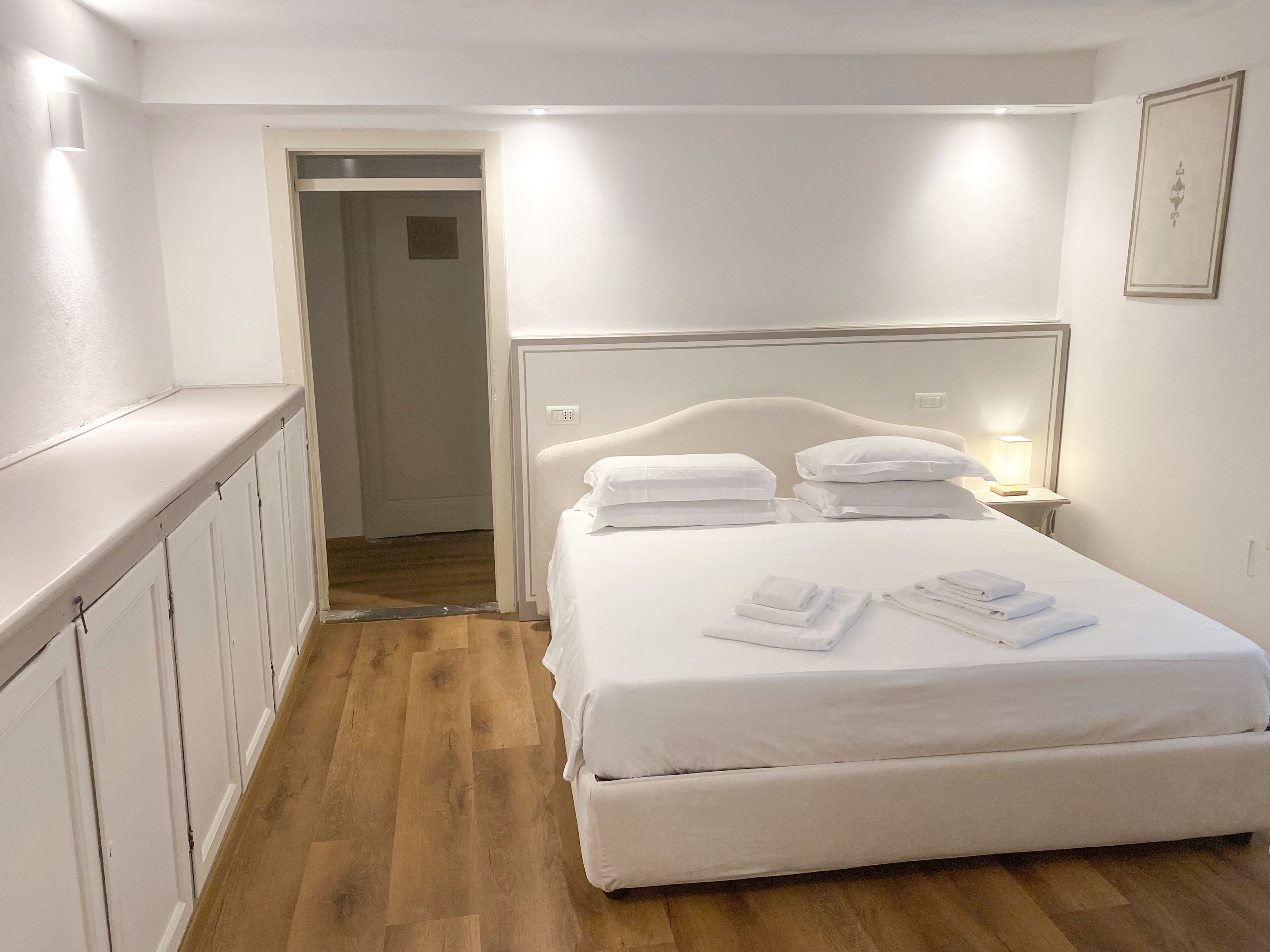 Una habitación de hotel con cama, mesita de noche, lámpara y una puerta que da a otra estancia.