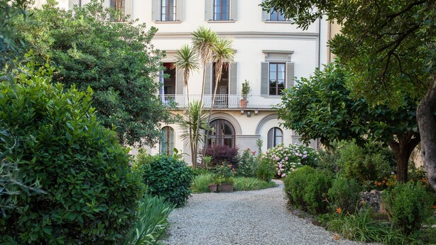 A gravel pathway leading to a building with a balcony, surrounded by lush greenery and various plants.