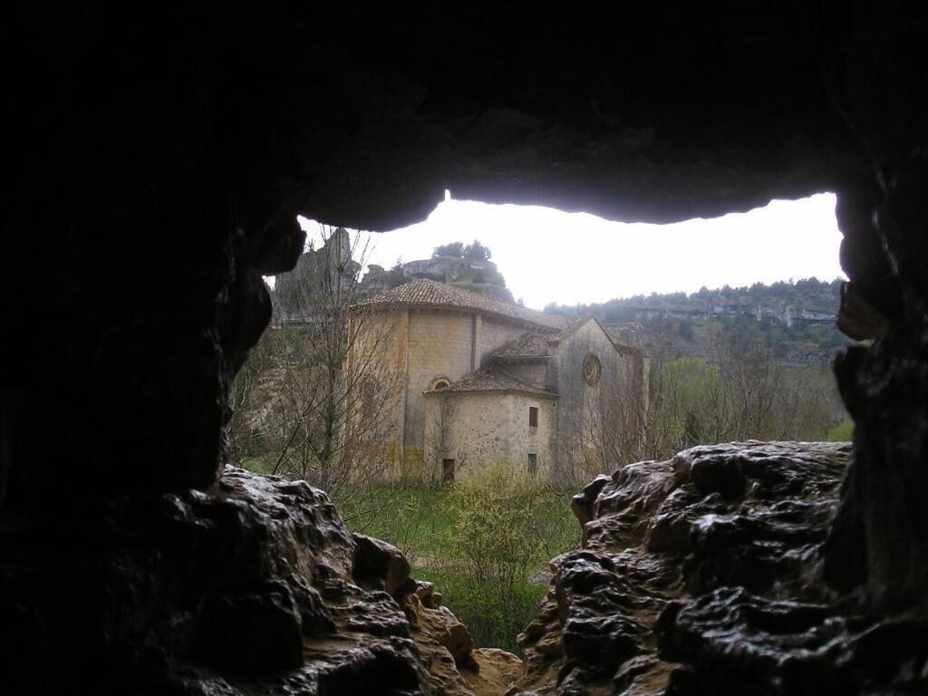 Vista a través de una abertura en la cueva, mostrando una construcción rústica con una cúpula y una edificación más pequeña, en un entorno natural.