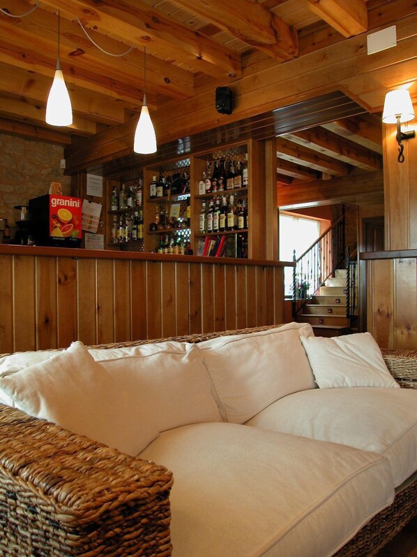 A cozy room with a wicker sofa, wooden ceiling beams, and a bar area with various bottles and a granini juice box.