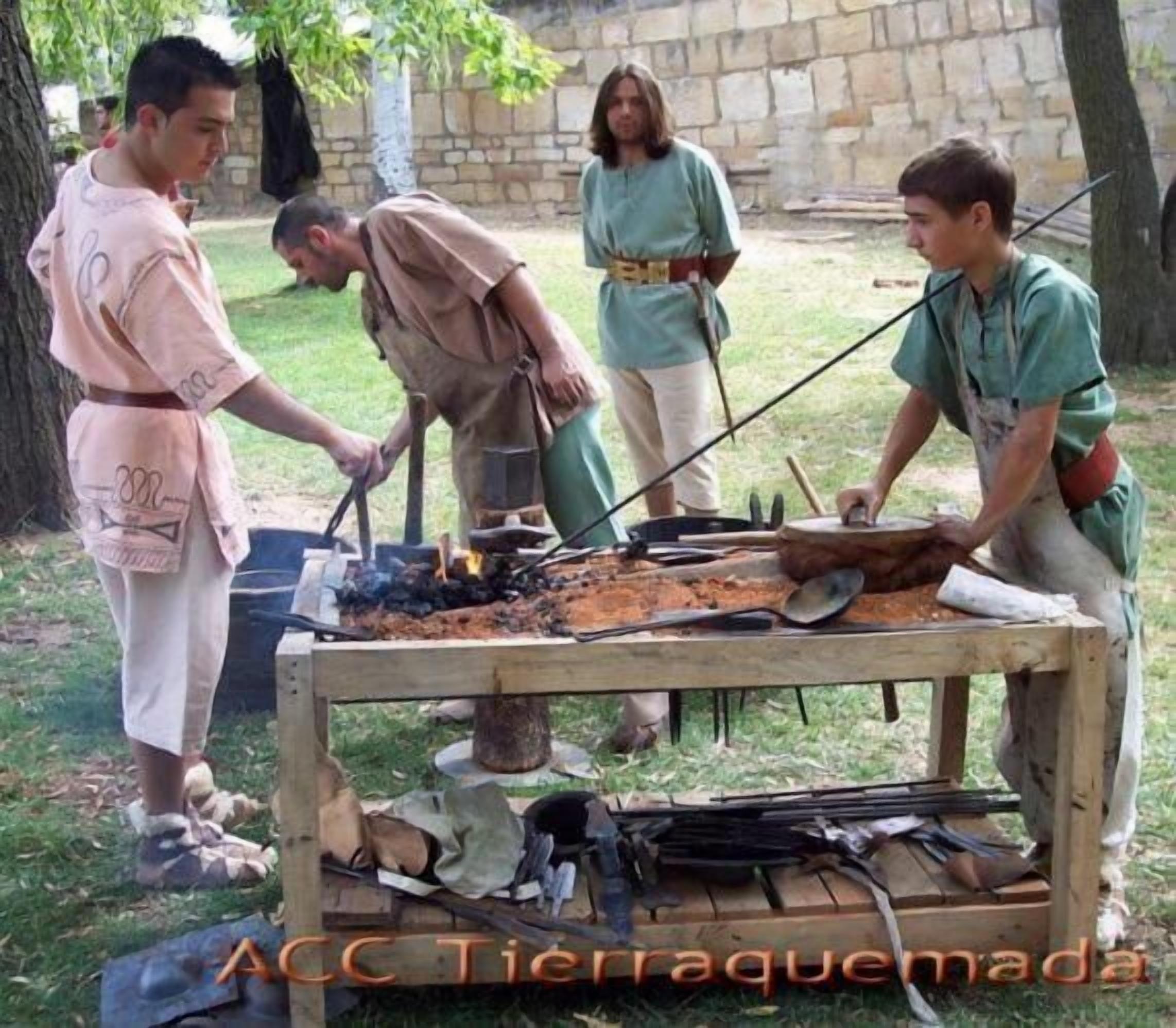 Un grup de persones participant en una activitat a l'aire lliure, possiblement una recreació històrica, amb una taula al centre que conté diverses eines i un foc.