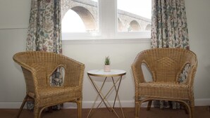 Two wicker chairs with a small round table between them, a potted plant on the table, and a view of a stone bridge through a window with floral curtains. - The Royal Oak Hotel (Buckie)