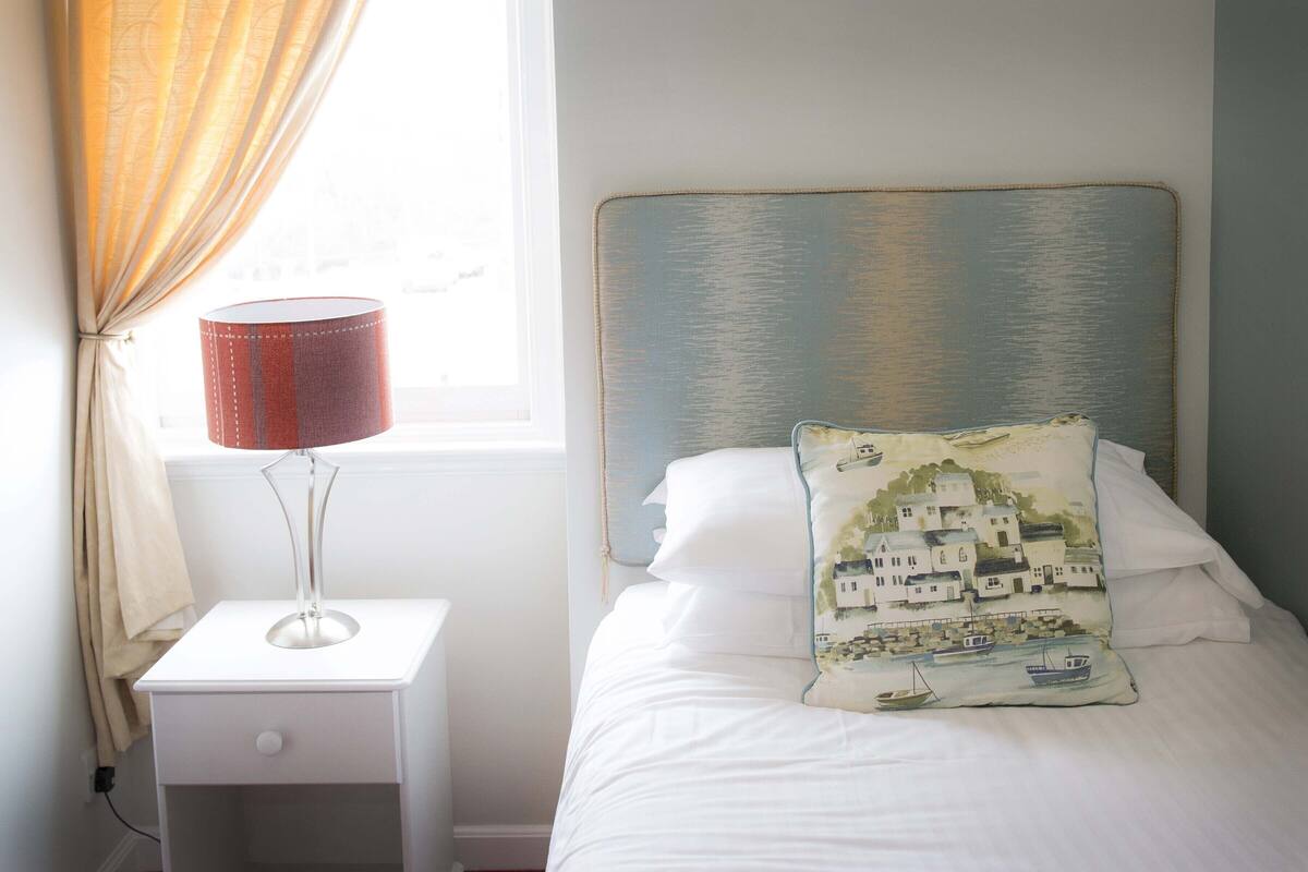 Una cama bien hecha con un almohadón decorativo, una lámpara de noche y una ventana con cortinas.
