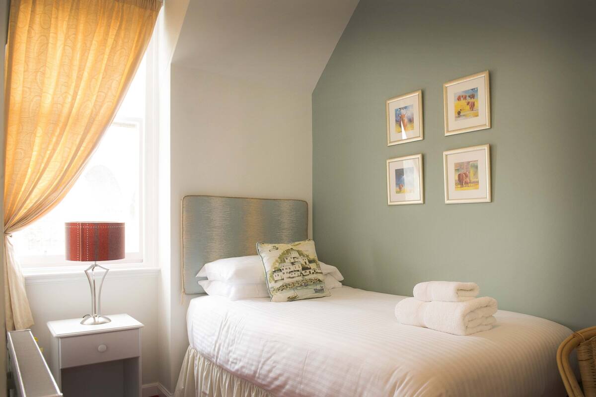 Una cama bien tendida con sábanas blancas, una lámpara de noche y una ventana con cortinas.