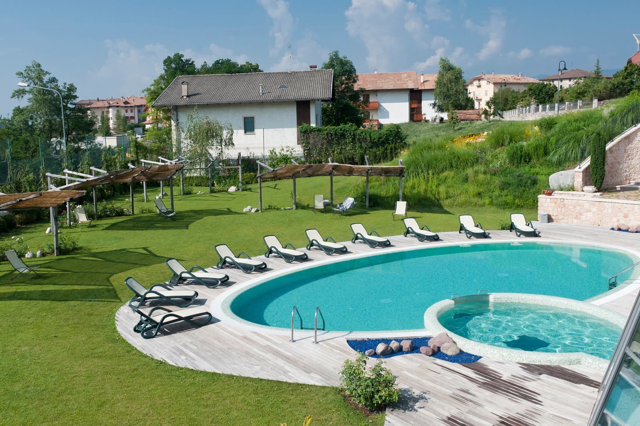 A swimming pool with lounge chairs and a hot tub, surrounded by a grassy area and a stone wall.