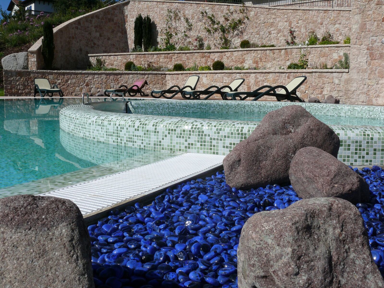 Ein Pool mit Mosaikfliesen, Liegestühlen und großen Steinen.