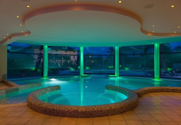 Una piscina interior moderna con un borde de mosaicos, ventanas amplias e iluminación ambiental.