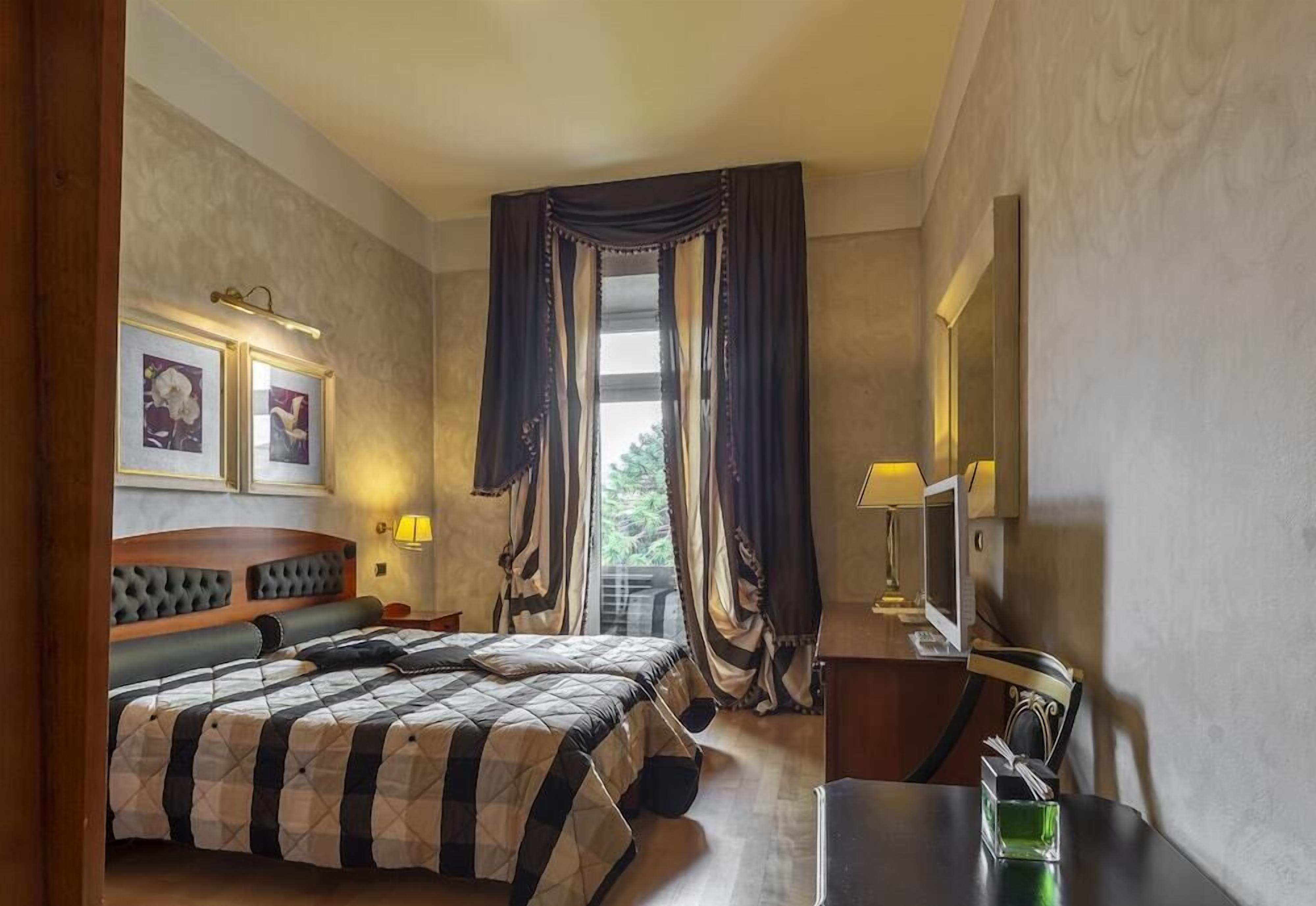 A hotel room with a large bed, a desk with a lamp, a television, and a window with curtains.