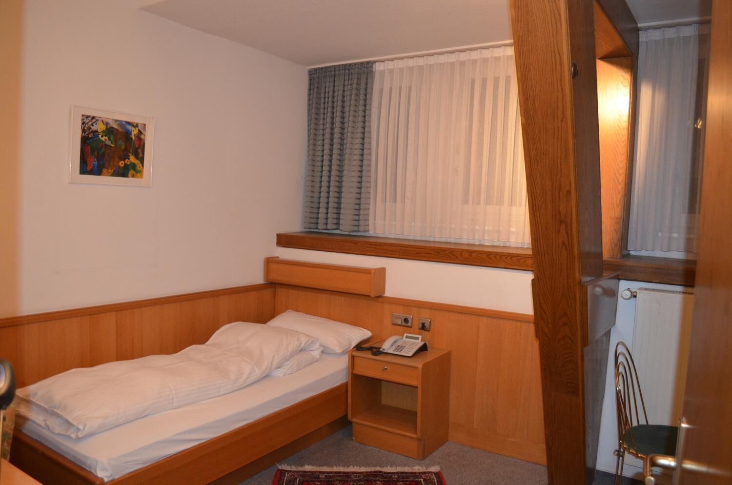 Une petite chambre avec un lit simple, une tête de lit en bois, une table de chevet avec un téléphone et une fenêtre avec des rideaux.