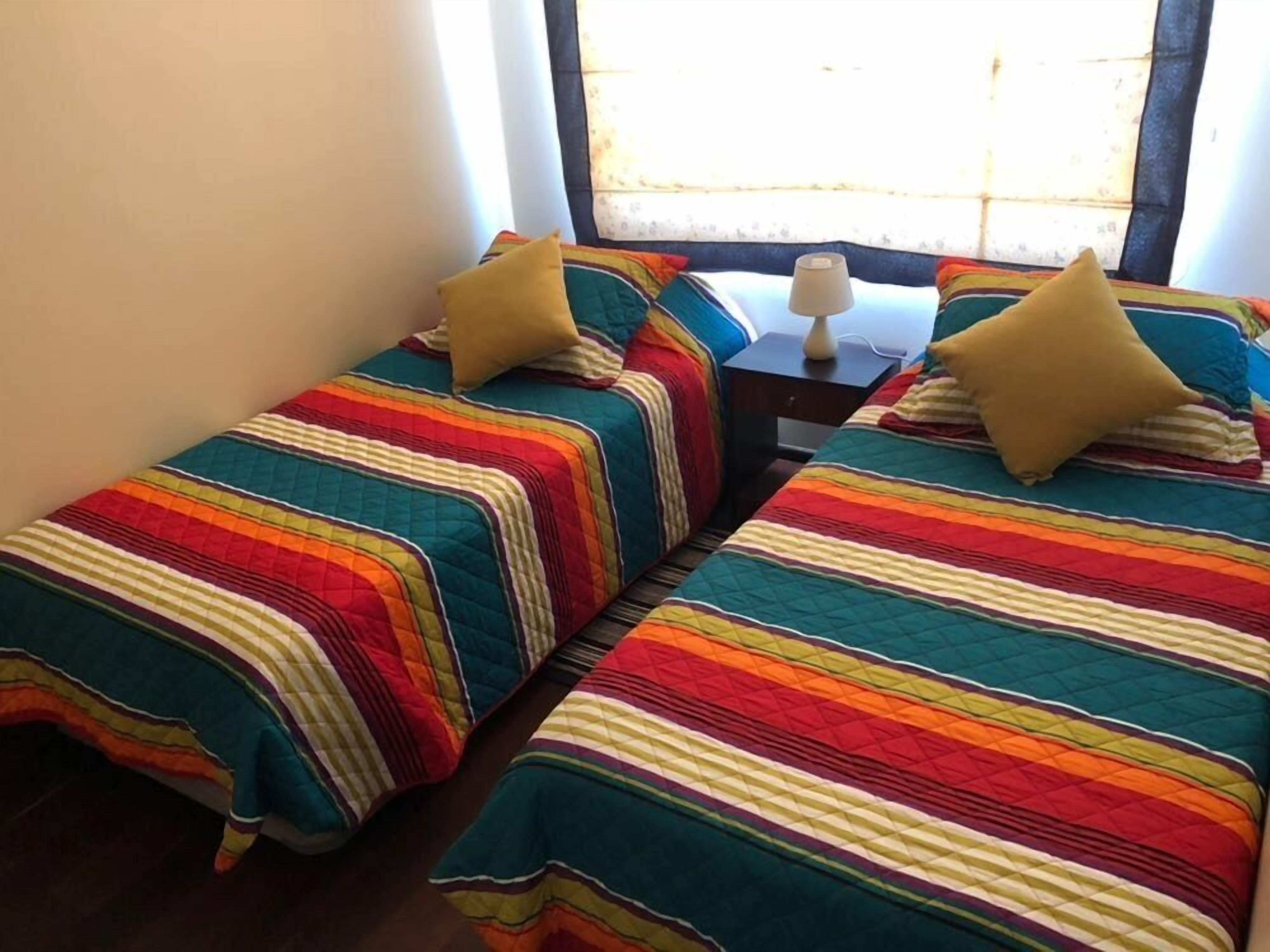 Una habitación con dos camas, cada una con una colcha a rayas colorida y una sola almohada. Hay una pequeña mesa con una lámpara entre las camas.