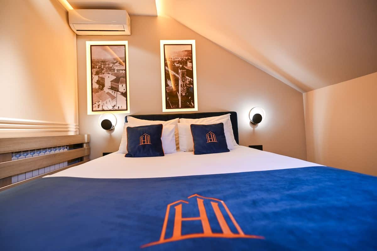 A hotel room with a bed, two framed pictures on the wall, and a logo on the bedspread.