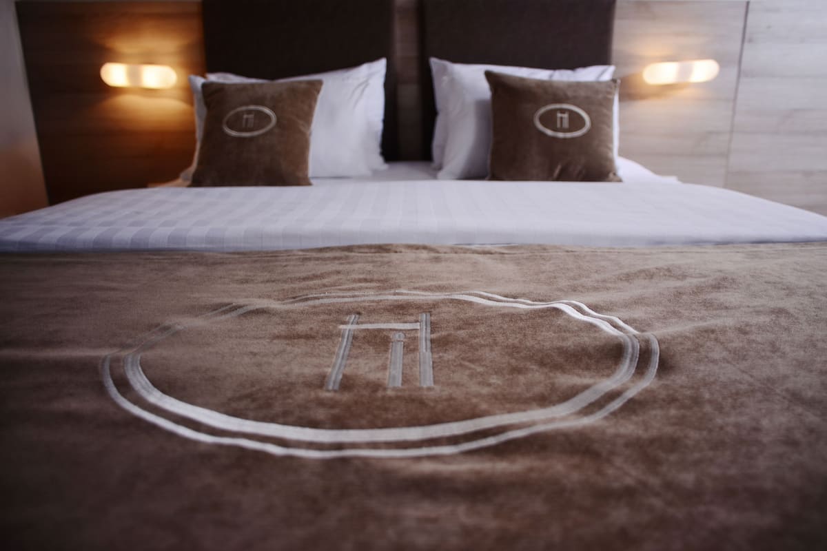 A bed with a brown blanket and pillows, a headboard with wall-mounted lights, and a visible logo on the pillows.
