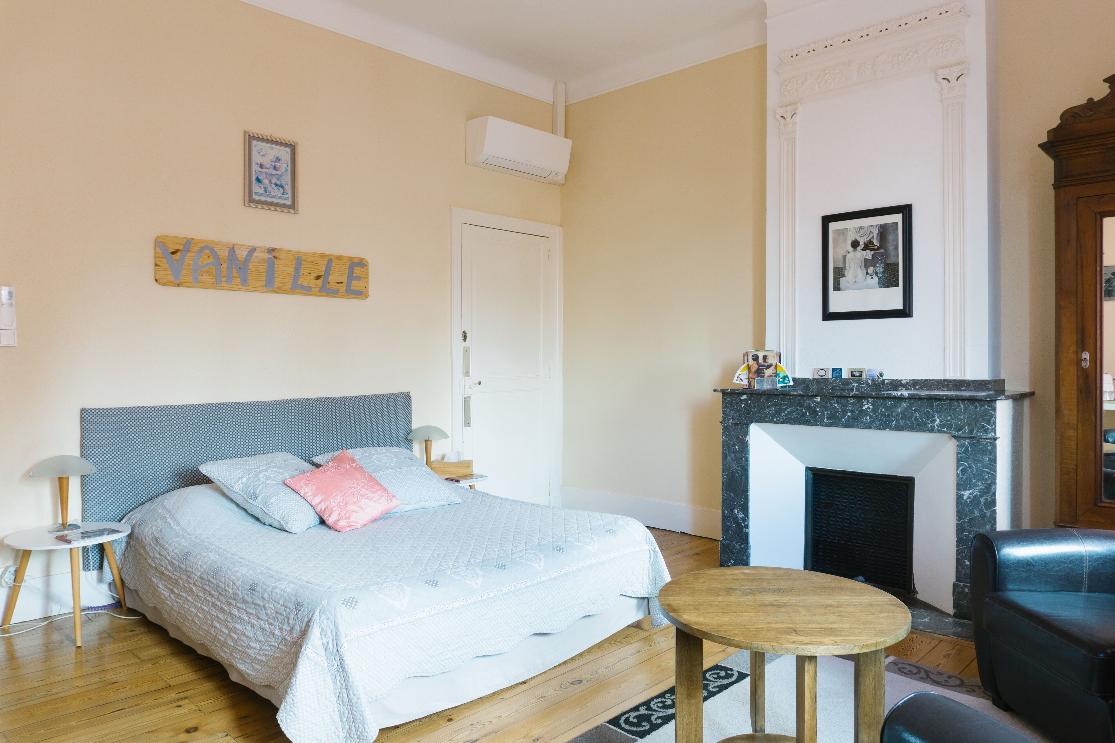 A bedroom with a bed, a small round table, a chair, a fireplace, and a wall-mounted air conditioner.
