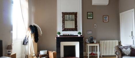 A hotel room with a fireplace, a bed, a small table, a radiator, and a window with curtains.