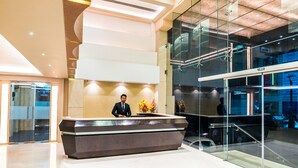 Zona de recepción moderna con un mostrador, un hombre de pie detrás y un muro de cristal que refleja el interior.