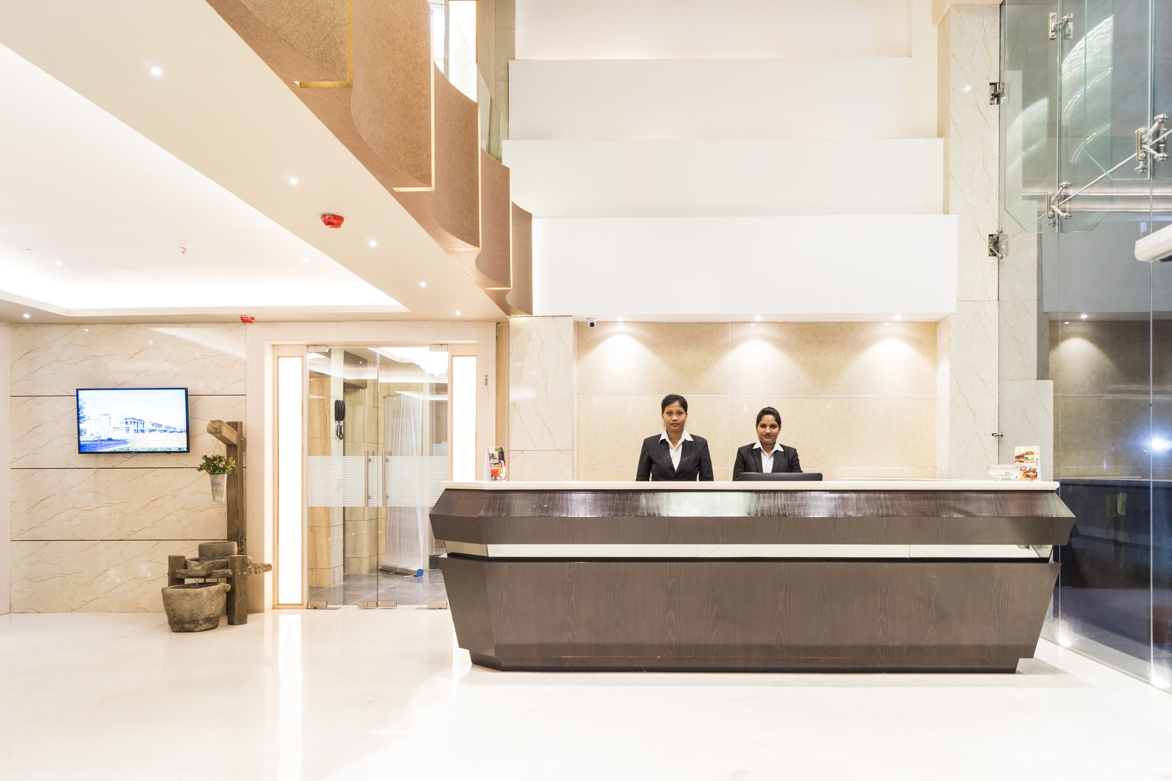 A reception desk with two staff members, a marble wall, a television, and a modern staircase.