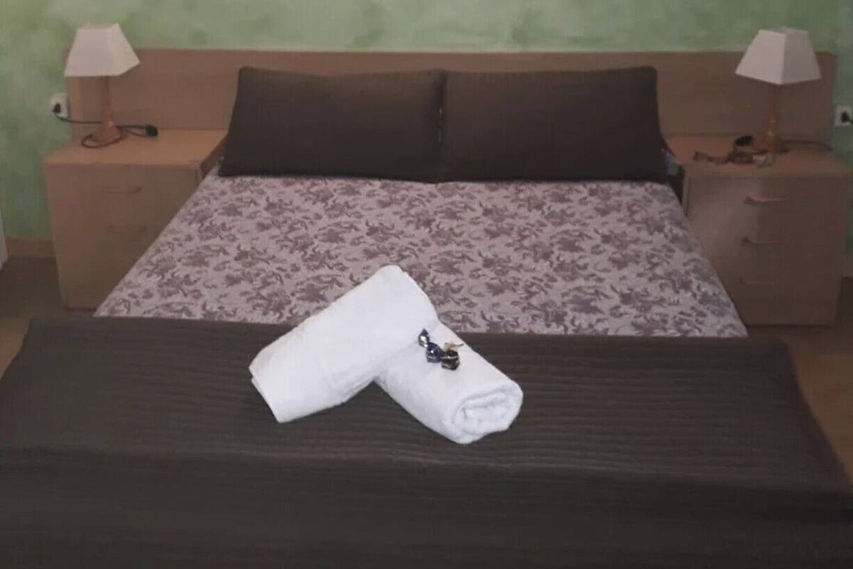 A neatly made bed with a floral bedspread, two pillows, and a folded towel on a dark headboard. There are two bedside tables with lamps and a wall-mounted light fixture above the bed.