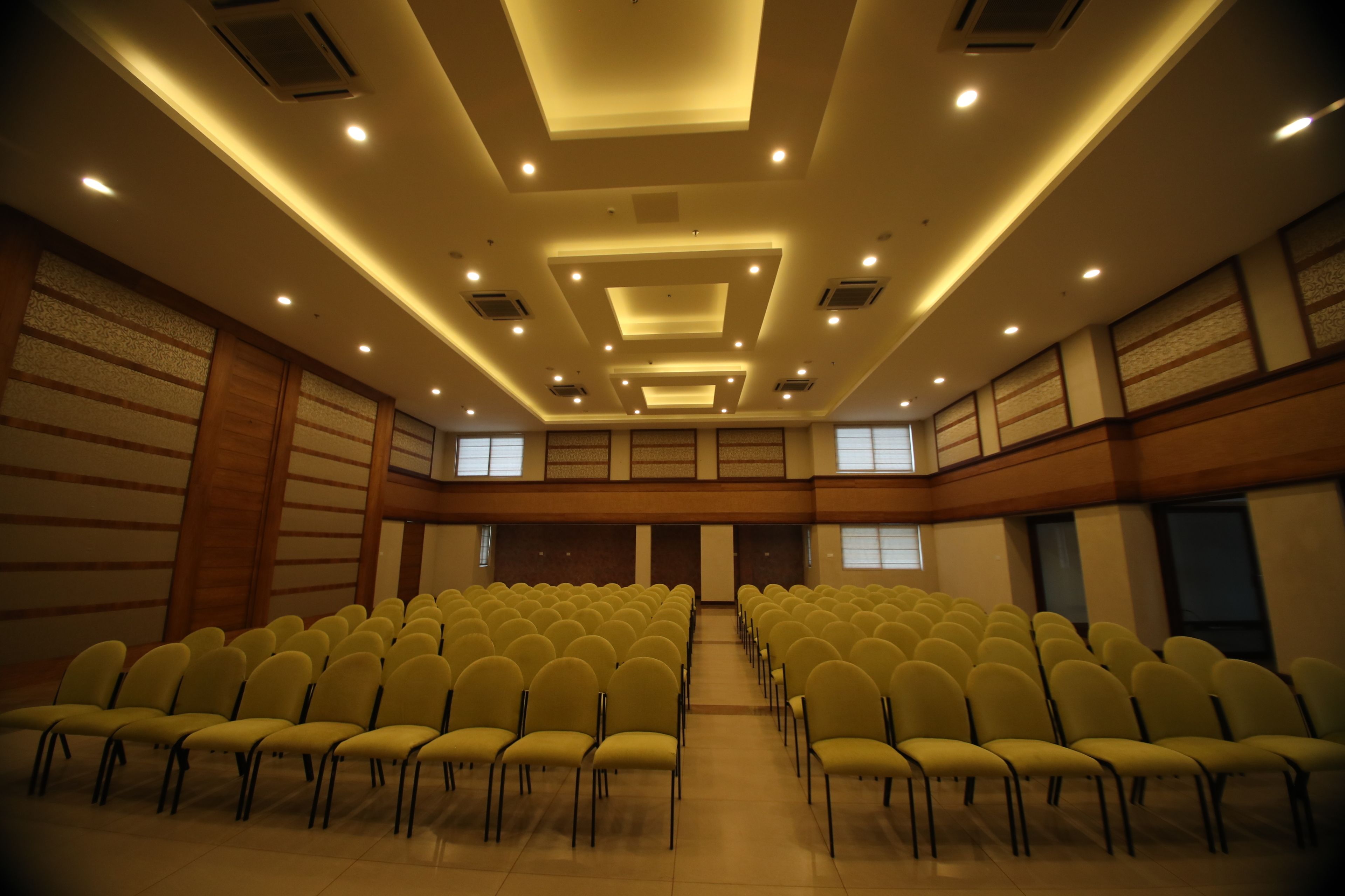 Un salón de conferencias con filas de sillas amarillas, un techo alto con iluminación empotrada y amplios ventanales a lo largo de la pared trasera.