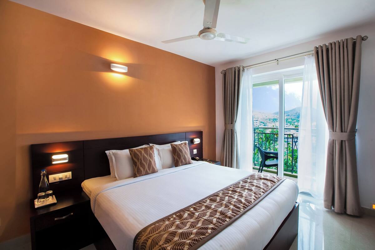 Une chambre d’hôtel avec un grand lit, une table de chevet avec une lampe, un ventilateur de plafond et une vue sur la verdure par la fenêtre.