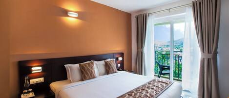 Habitación de hotel con una cama grande, una mesita de noche con lámpara, un ventilador de techo y vistas a zonas verdes por la ventana.
