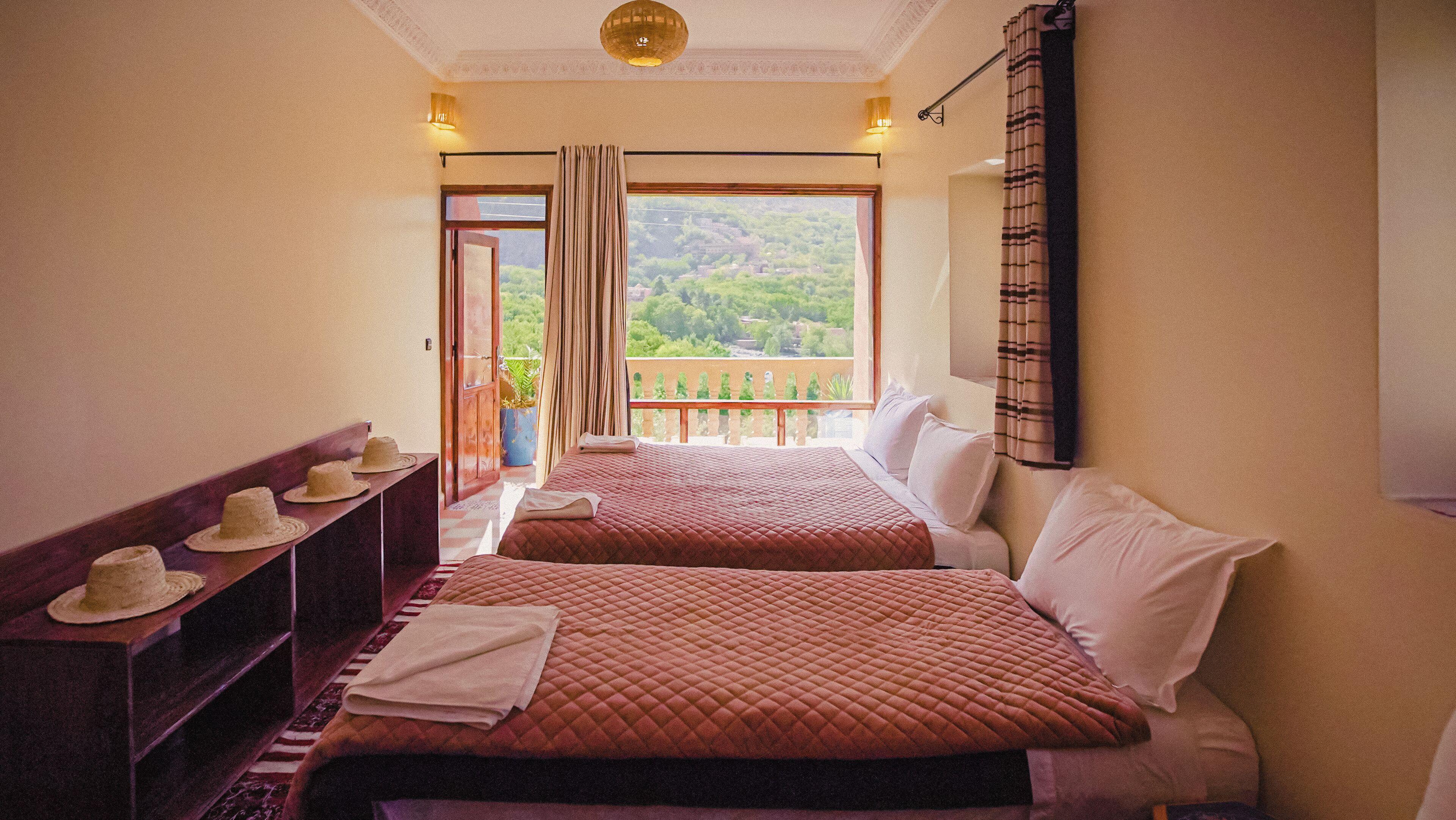 A hotel room with two beds, a balcony with a view of greenery, and a small table with hats.
