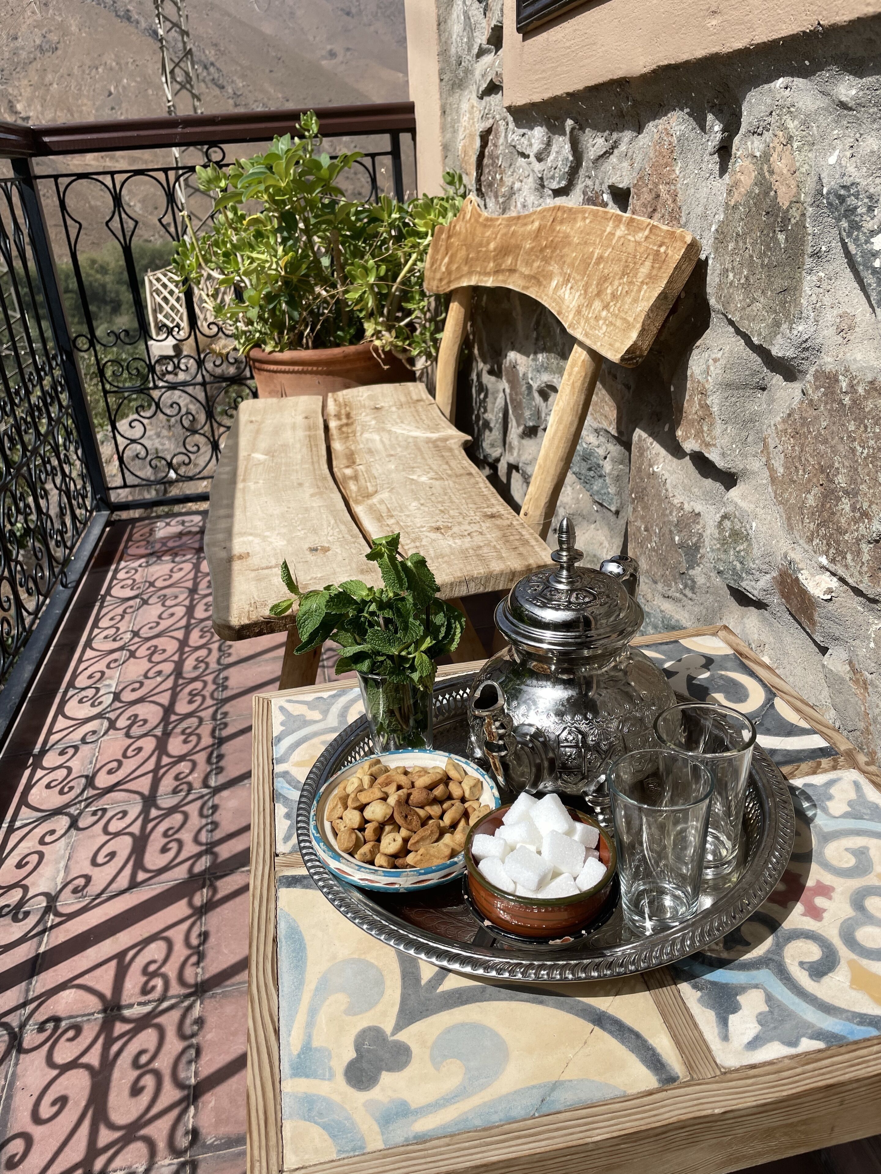 A table set with a teapot, sugar cubes, and nuts on a patterned tablecloth, with a potted plant and a wooden chair in the background.