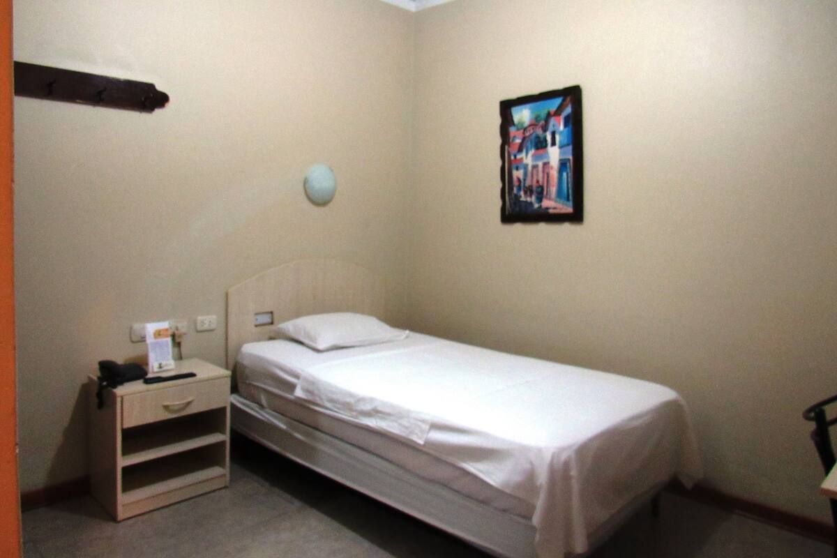 A small, single-bed room with a wooden nightstand, a wall-mounted light, and a framed picture.