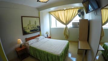 Quarto de hotel com cama, mesa de cabeceira, luminária, televisão e janela com cortinas.