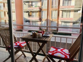 Un balcón con una mesa de madera y dos sillas, vista a un edificio de varios pisos con balcones, y una barandilla.