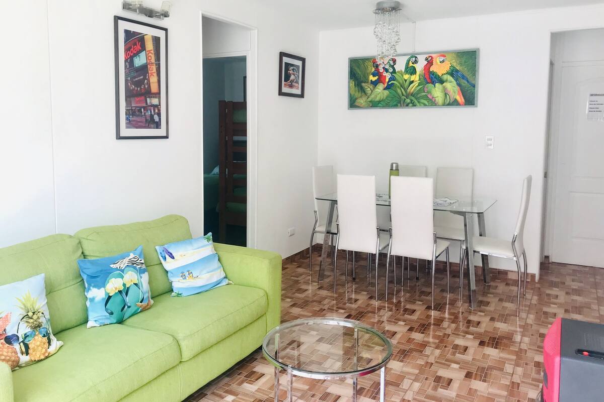 A living room with a green sofa, a glass coffee table, white chairs, a dining table, and a painting of parrots.