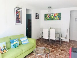 Un salón con un sofá verde, una mesa de centro de vidrio, sillas blancas, una mesa de comedor y un cuadro de loros.