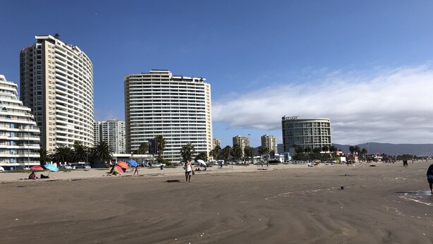 Una playa con edificios altos al fondo, incluyendo un hotel con un cartel visible.