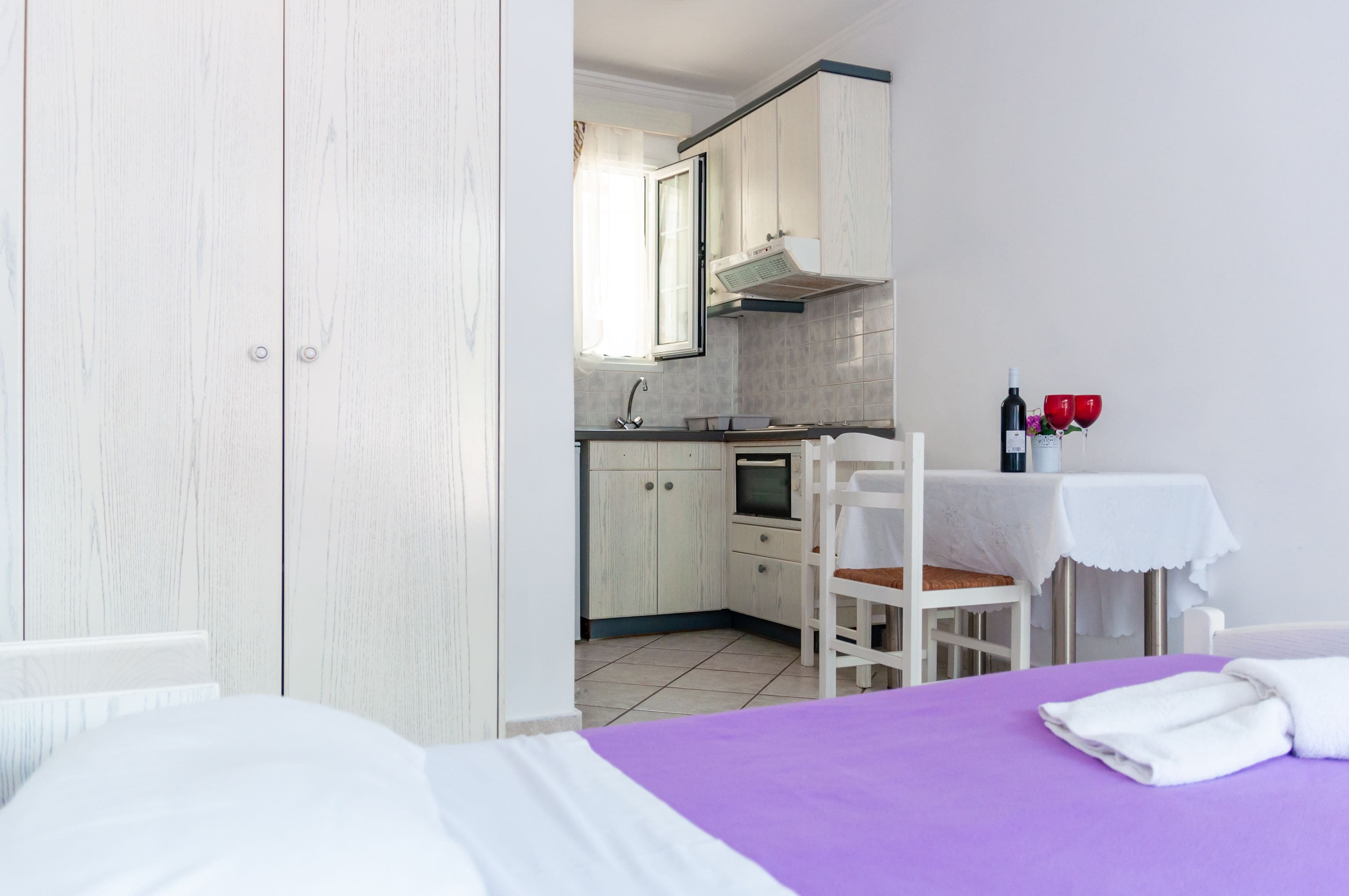 A room with a bed, a purple bedspread, a white wardrobe, a kitchen area with cabinets, a window, and a table with a bottle and glasses.
