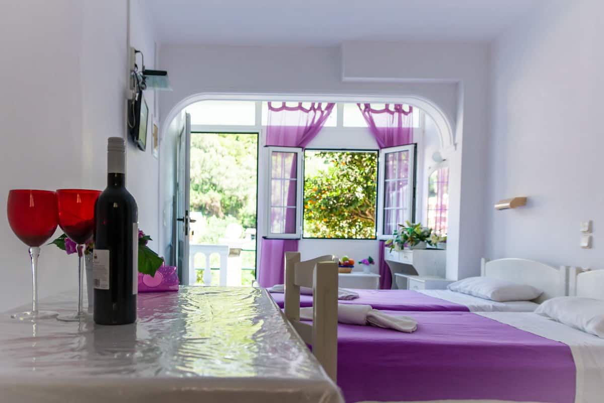 A room with two beds, a table with a bottle and glasses, and a view of greenery through open windows.