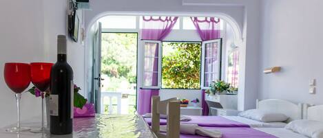 A room with two beds, a table with a bottle and glasses, and a view of greenery through open windows.