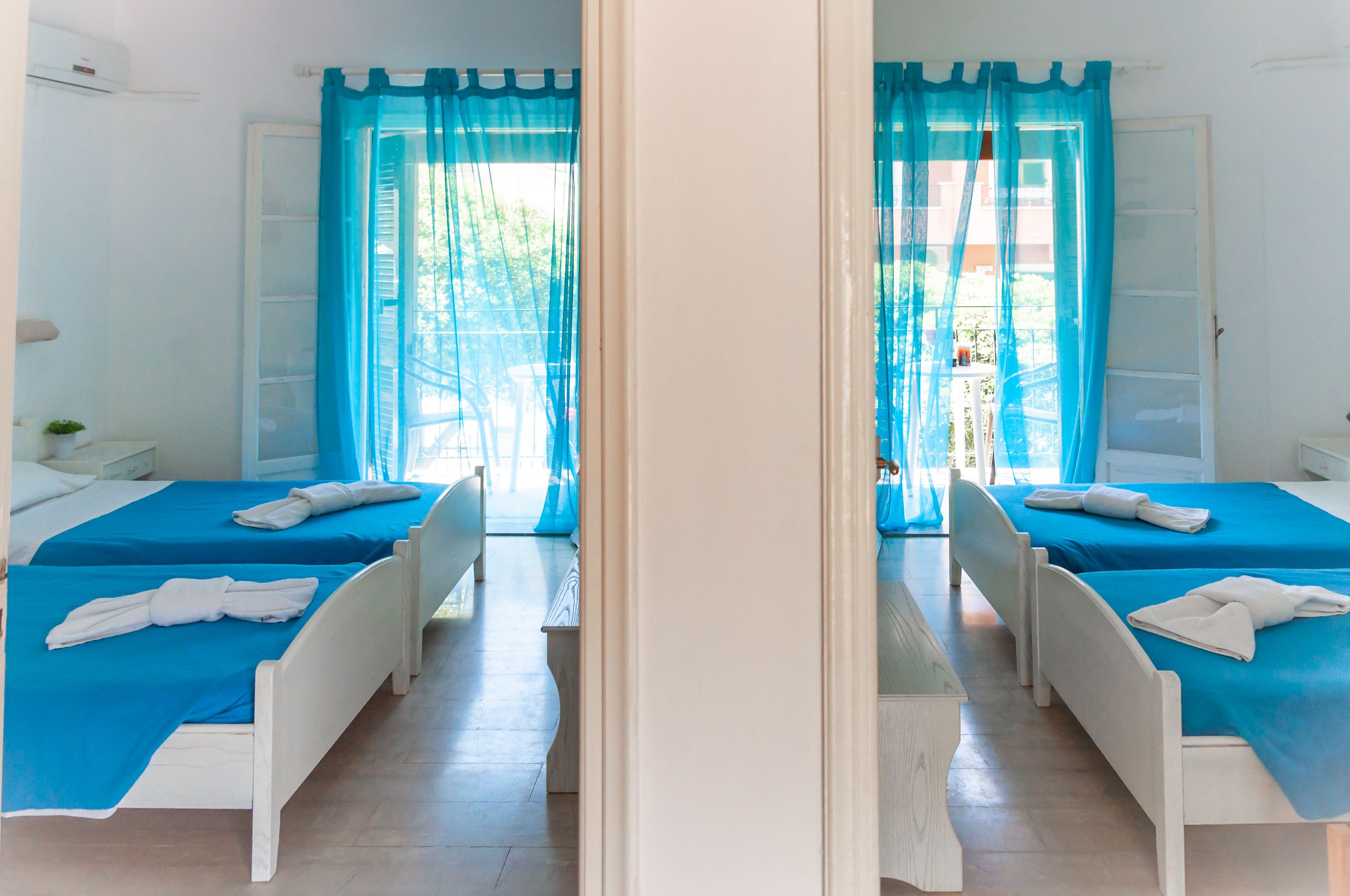 Reflejo de una habitación de hotel con dos camas, cada una con colcha azul y cabeceros blancos, y un ventanal con cortinas azules.