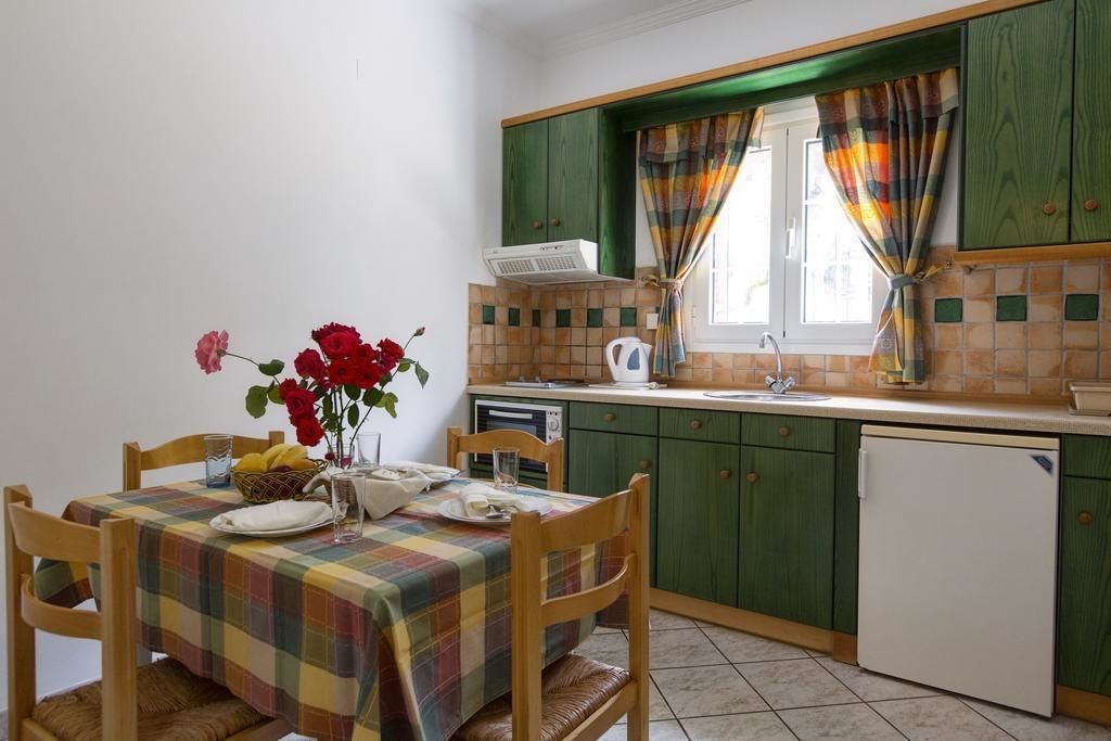 Una cucina con mobili verdi, zoccolo a mosaico, finestra con tende, tavolo da pranzo con tovaglia a quadri e un vaso di fiori.