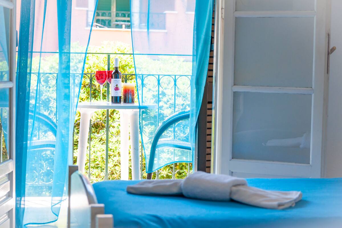 A room with a bed, a window with blue curtains, and a small table with bottles and a red flower.