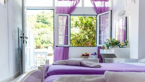 Un dormitorio con una cama, un ventanal con cortinas moradas, una mesita de noche con una cesta de fruta y vistas al verdor exterior.