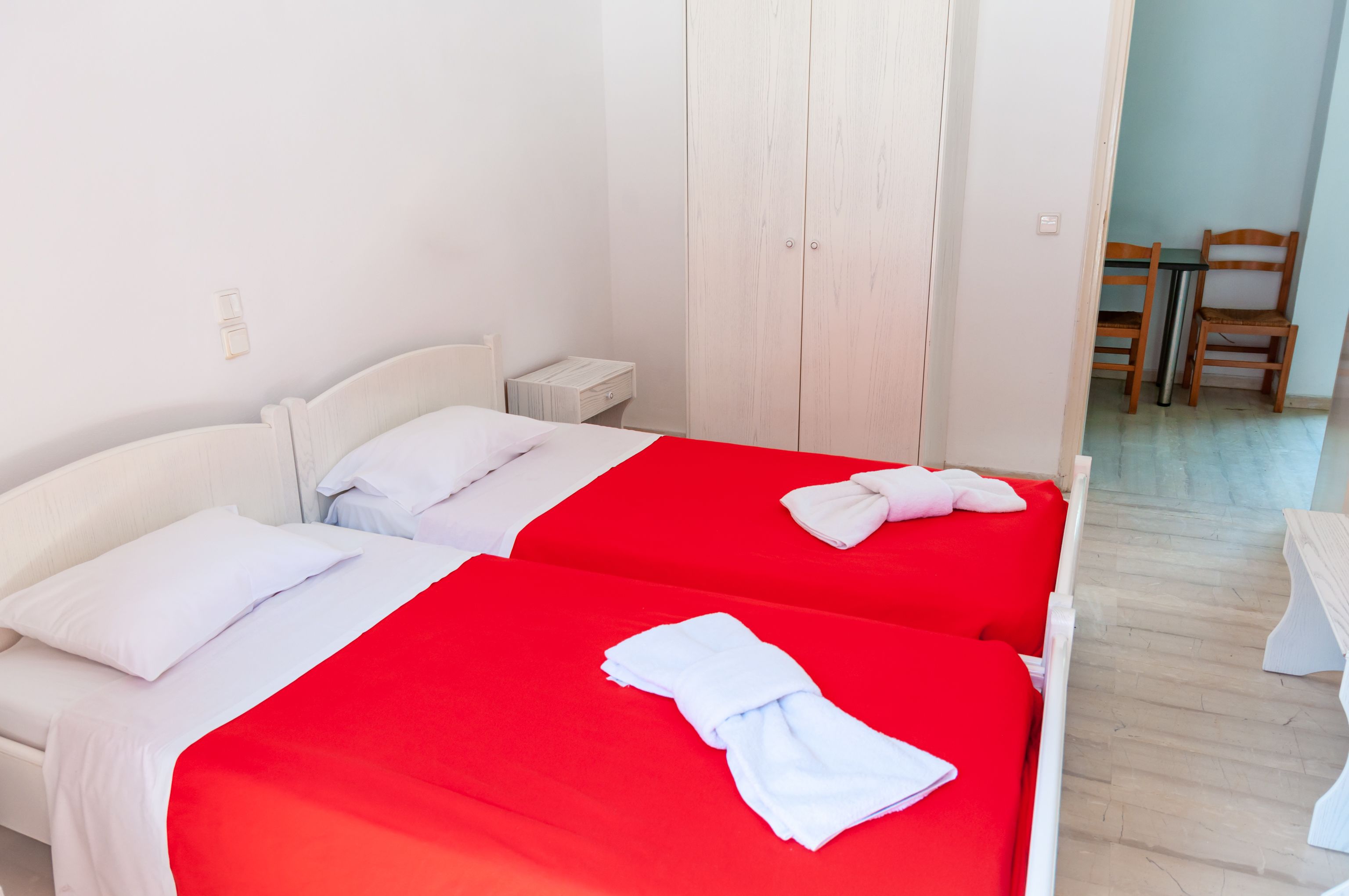 Habitación de hotel con dos camas, cada una con cubrecama rojo y almohadas blancas. Se observa un armario de madera, una mesita de noche pequeña y una silla en la esquina.